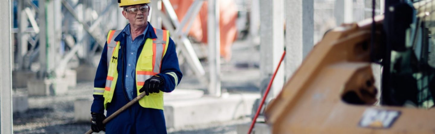 A construction worker wearing a hard hat, safety glasses, and a reflective vest uses a rake at an industrial site with metal structures and equipment visible in the background.