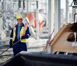 A construction worker wearing a hard hat, safety glasses, and a reflective vest uses a rake at an industrial site with metal structures and equipment visible in the background.