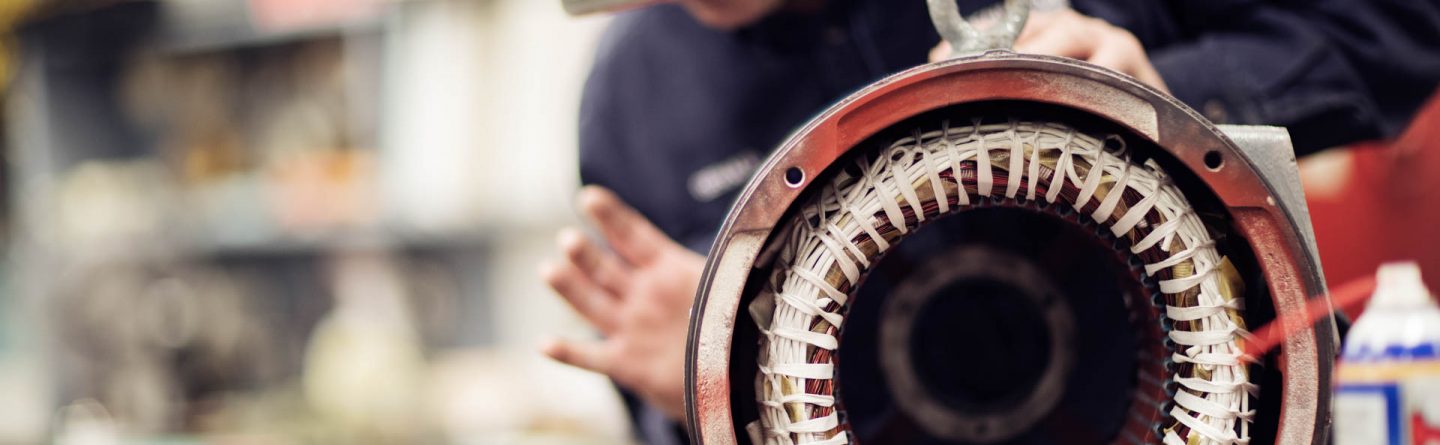 A person wearing a cap works on an industrial electric motor, focusing on the exposed wiring and inner components in a workshop setting. The motor’s coils are clearly visible in the foreground.