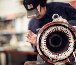 A person wearing a cap works on an industrial electric motor, focusing on the exposed wiring and inner components in a workshop setting. The motor’s coils are clearly visible in the foreground.