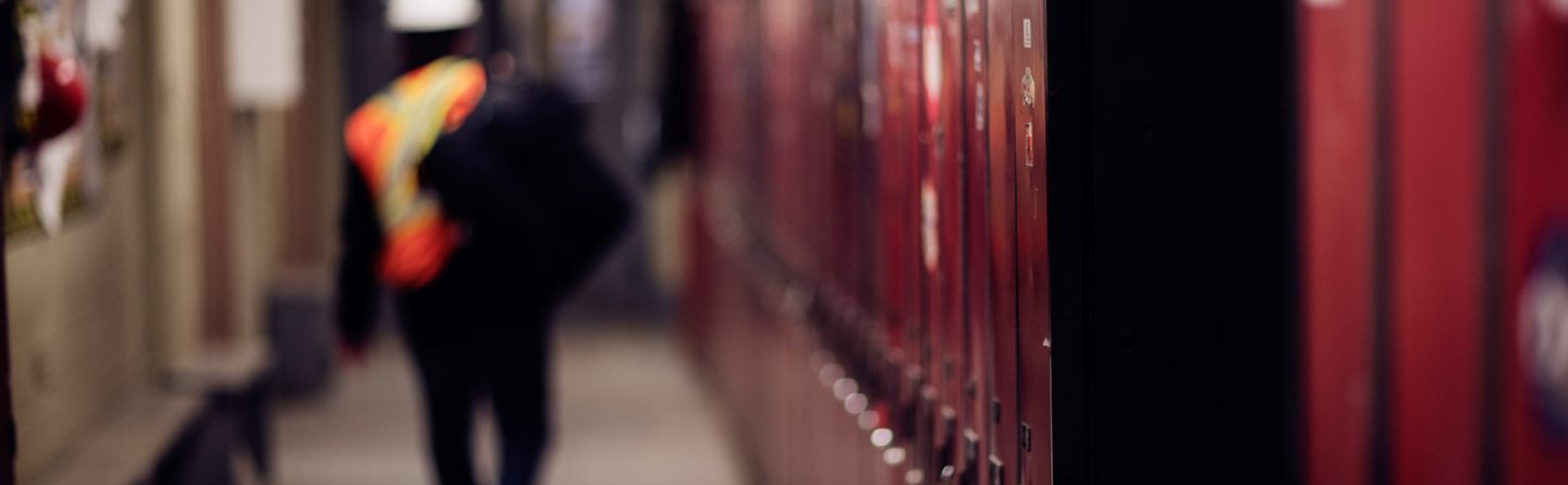 A worker in a reflective vest and hard hat walks down a hallway lined with red lockers. The image is focused on the lockers, with the worker blurred in the background.