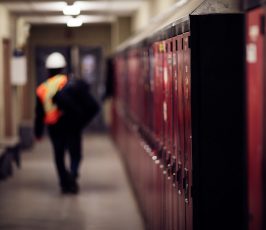 A worker in a reflective vest and hard hat walks down a hallway lined with red lockers. The image is focused on the lockers, with the worker blurred in the background.