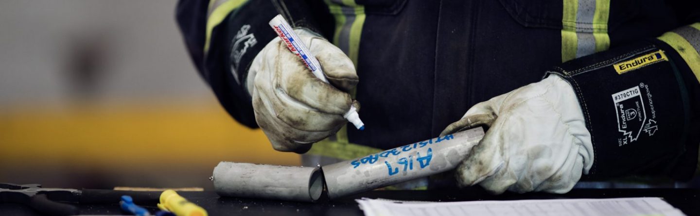 A person wearing protective gloves and reflective work clothing writes on a metal pipe with a marker, with tools and a document on a table in front of them.
