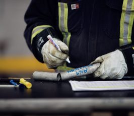 A person wearing protective gloves and reflective work clothing writes on a metal pipe with a marker, with tools and a document on a table in front of them.