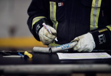A person wearing protective gloves and reflective work clothing writes on a metal pipe with a marker, with tools and a document on a table in front of them.