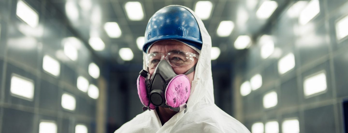 A person wearing a blue hard hat, safety goggles, a white protective suit, and a respirator with pink filters stands in a brightly lit industrial facility.
