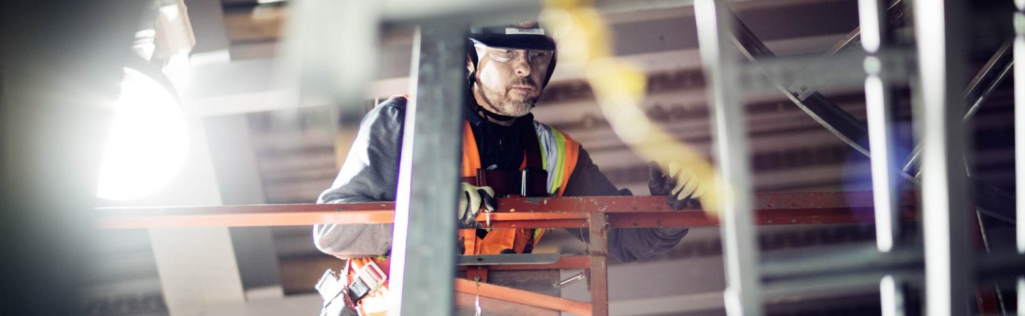 A construction worker wearing a safety vest and helmet stands on a lift, working indoors near metal beams and industrial lighting. The scene is faintly illuminated, with equipment visible in the foreground.