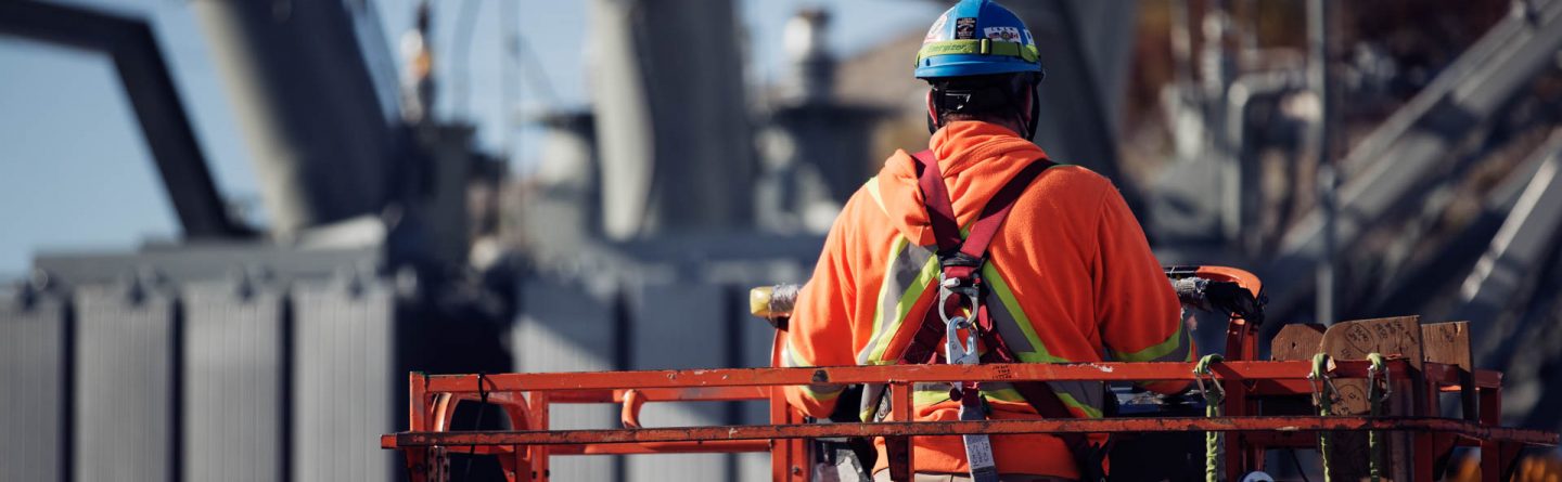 A construction worker wearing a hard hat and orange safety jacket stands on a lift platform at an industrial site with large electrical equipment in the background.