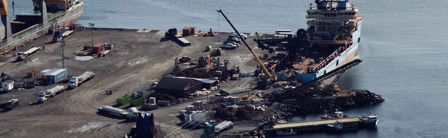 Aerial view of a dock with two large cargo ships moored alongside. Cranes, vehicles, and various equipment are scattered across the pier, with calm water surrounding the area.