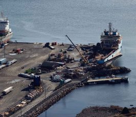 Aerial view of a dock with two large cargo ships moored alongside. Cranes, vehicles, and various equipment are scattered across the pier, with calm water surrounding the area.