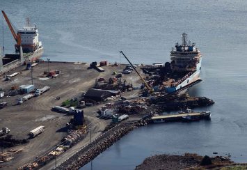 Aerial view of a dock with two large cargo ships moored alongside. Cranes, vehicles, and various equipment are scattered across the pier, with calm water surrounding the area.