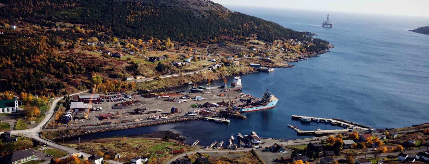 Aerial view of a coastal village with docks, ships, scattered houses, and autumn-colored trees, surrounded by hills and bordered by a calm blue sea with an offshore oil platform visible in the distance.