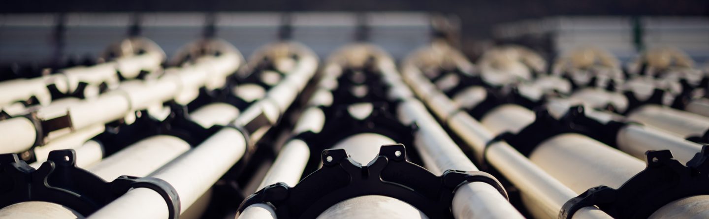 Close-up view of parallel white industrial pipes secured with black brackets, stretching into the distance with a blurred landscape and sky in the background.