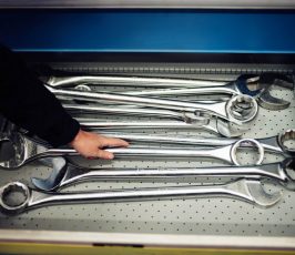 A person’s hand reaching for a large metal wrench among several oversized wrenches arranged in a drawer with a perforated metal bottom.