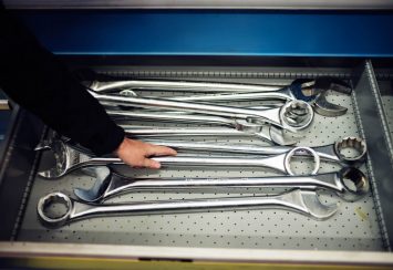 A person’s hand reaching for a large metal wrench among several oversized wrenches arranged in a drawer with a perforated metal bottom.