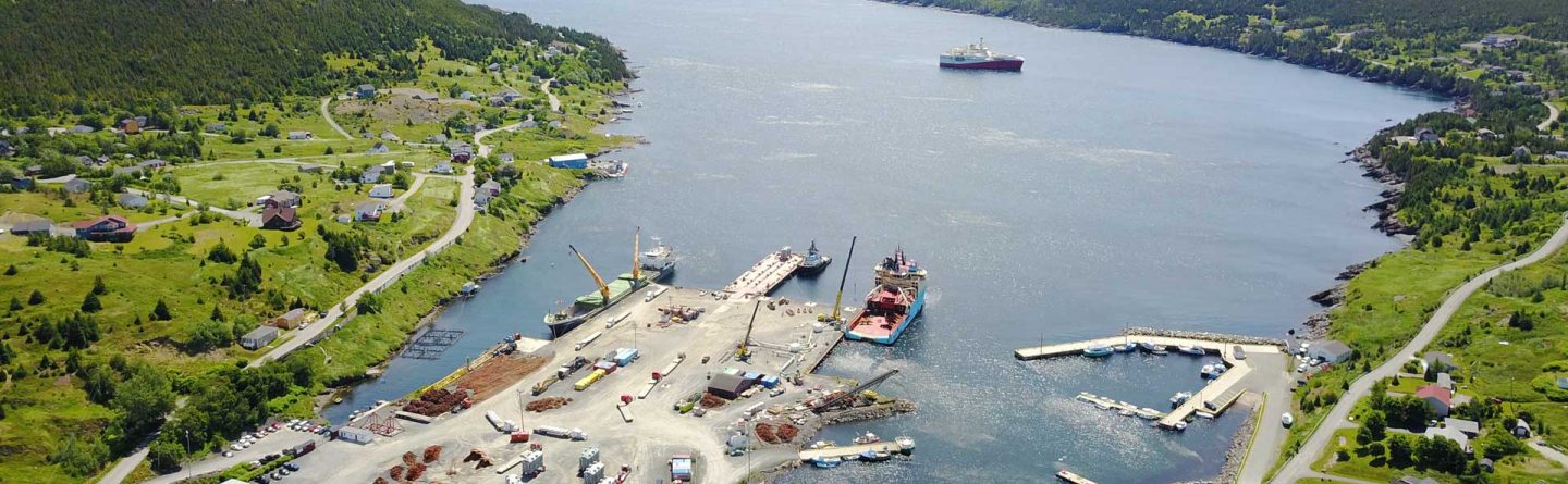 Aerial view of a harbor with several ships docked, surrounded by green hills and scattered buildings along the coastline, with open sea and ships in the distance under a clear sky.