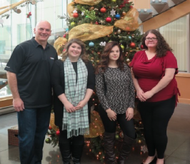 Four people stand smiling in front of a decorated Christmas tree indoors. Three women are dressed in winter clothes, and one man is in a black shirt. The tree is adorned with ornaments, ribbons, and lights.