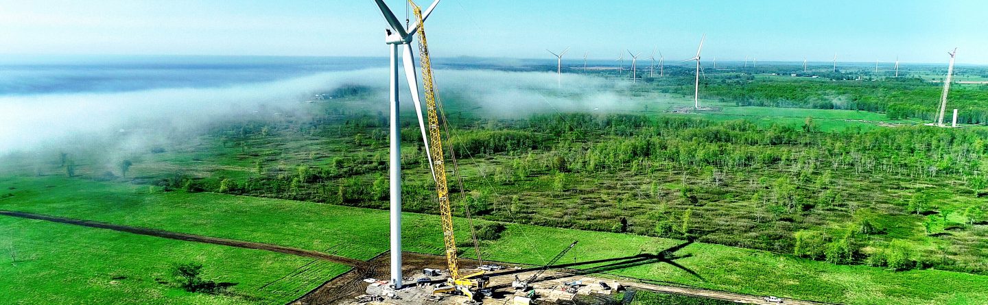 A large crane installs blades on a wind turbine in a green field, surrounded by construction equipment and workers. Other wind turbines and a low fog are visible in the distance under a clear blue sky.