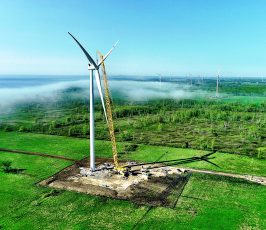 A large crane installs blades on a wind turbine in a green field, surrounded by construction equipment and workers. Other wind turbines and a low fog are visible in the distance under a clear blue sky.