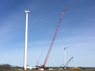 A large red crane lifts a wind turbine component next to a tall, incomplete wind turbine tower. Another wind turbine and crane are visible in the background under a clear blue sky.