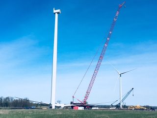 A large red crane lifts a wind turbine blade next to a tall white wind turbine tower. Another wind turbine and crane are visible in the background under a clear blue sky over a grassy field.