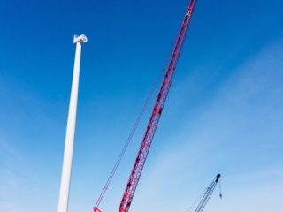 A large red crane lifts a component near a tall white wind turbine tower on a grassy field, with a smaller black crane and yellow equipment nearby under a clear blue sky.