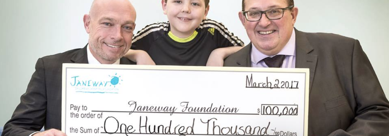 Three smiling people, two men in suits and a boy in a black shirt, hold a large ceremonial check for $100,000 made out to the Janeway Foundation, dated March 2, 2017.