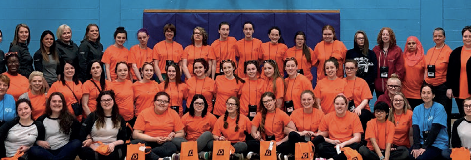 A large group of people, mostly women, pose indoors against a blue wall. Most are wearing matching orange shirts, while a few wear other colors. Some are seated at the front, others stand behind them, all smiling at the camera.