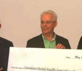Three people stand indoors holding a large ceremonial check for $250,000, addressed to the Canadian Mental Health Association NL, dated May 11, 2018. The check features the Pennecon logo. Two people wear green ribbons.