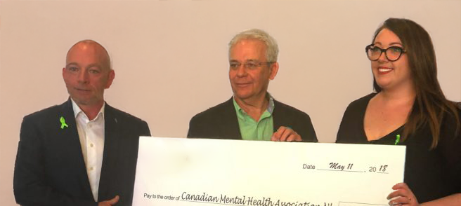 Three people stand indoors holding a large ceremonial check for $250,000, addressed to the Canadian Mental Health Association NL, dated May 11, 2018. The check features the Pennecon logo. Two people wear green ribbons.