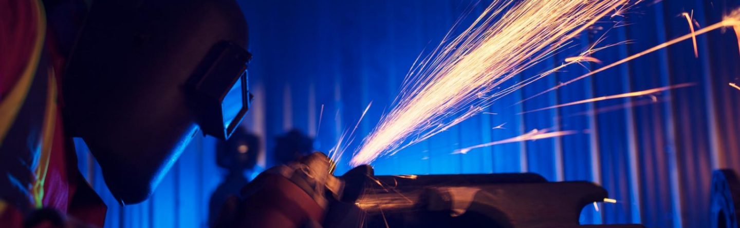A worker wearing a protective welding helmet and reflective vest uses a grinder on metal, creating bright orange sparks in a dimly lit industrial setting with blue lighting.
