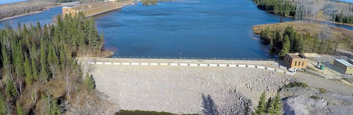Aerial view of a dam holding back a large body of water, surrounded by trees and a forested landscape under a bright blue sky with scattered clouds. Buildings are visible near the water’s edge.