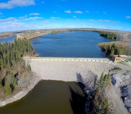 Aerial view of a dam holding back a large body of water, surrounded by trees and a forested landscape under a bright blue sky with scattered clouds. Buildings are visible near the water’s edge.