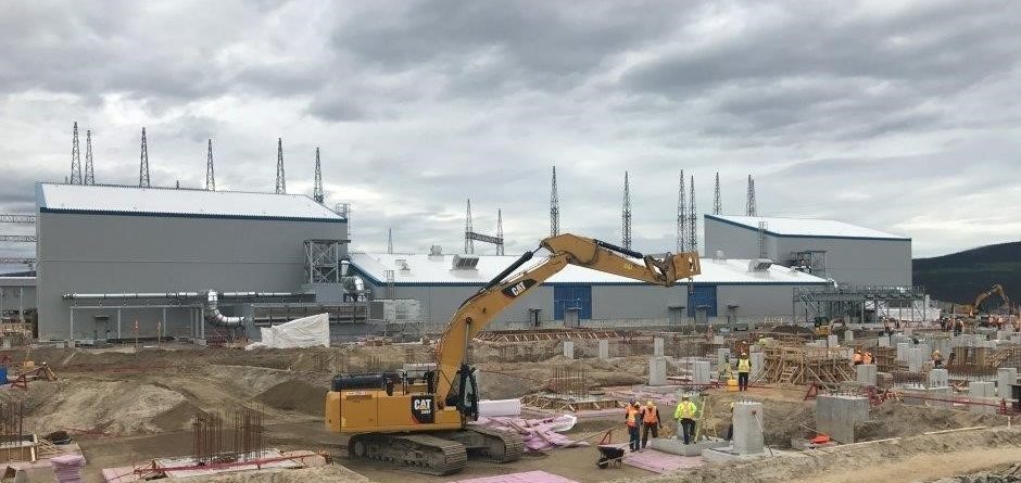 A construction site with workers in safety gear and a yellow excavator. Industrial buildings with metal roofs are in the background under a cloudy sky, and concrete foundations are being prepared in the foreground.