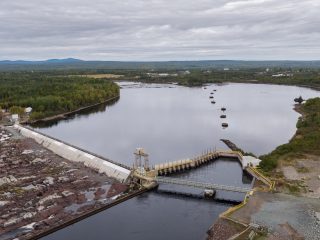 Aerial view of a hydroelectric dam with water flowing through a spillway, surrounded by rocky terrain, trees, and a large, calm river under a cloudy sky.