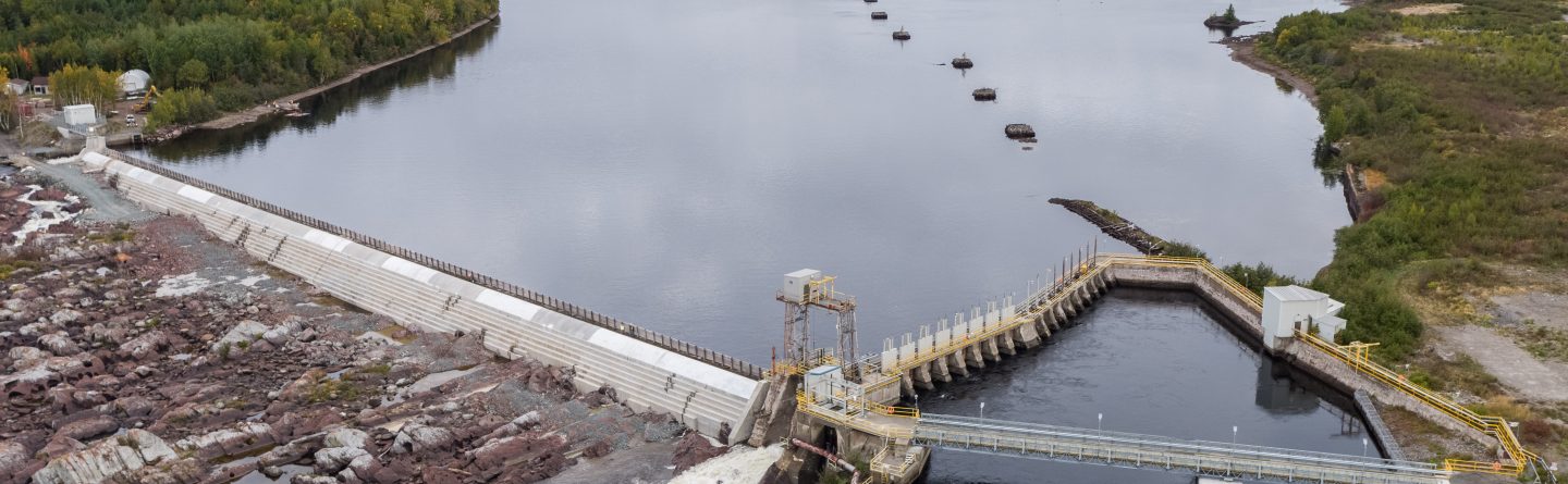 Aerial view of a hydroelectric dam spanning a wide river, with rocky terrain on one side and lush green trees and fields in the background under a cloudy sky.