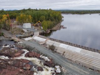 An aerial view of a dam with water flowing over its edge, surrounded by trees with some autumn foliage. Construction equipment and a small building are visible near the dam, and a large river extends into the distance.