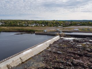 A large concrete dam stretches across a river, with water on one side and rocky terrain on the other. In the background, there is a small town surrounded by trees under a cloudy sky.