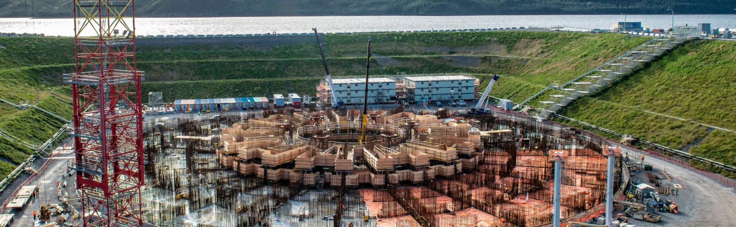 Aerial view of a large circular construction site with scaffolding, cranes, and building materials, surrounded by green hills and a body of water in the background under a cloudy sky.
