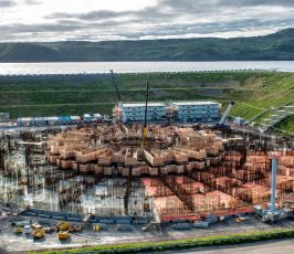 Aerial view of a large circular construction site with scaffolding, cranes, and building materials, surrounded by green hills and a body of water in the background under a cloudy sky.