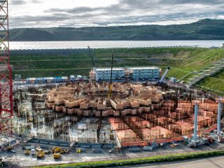 Aerial view of a large circular construction site with scaffolding, cranes, and building materials, surrounded by green hills and a body of water in the background under a cloudy sky.