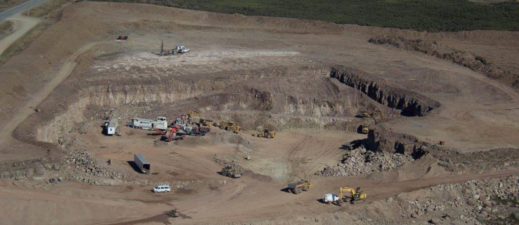 Aerial view of an open-pit mining site with large excavators, trucks, and construction equipment working on exposed earth surrounded by rocky terrain and sparse vegetation. A road runs along the top left of the site.