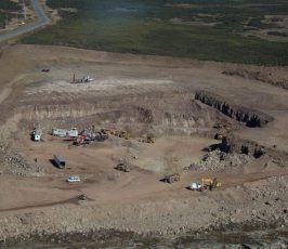 Aerial view of an open-pit mining site with large excavators, trucks, and construction equipment working on exposed earth surrounded by rocky terrain and sparse vegetation. A road runs along the top left of the site.