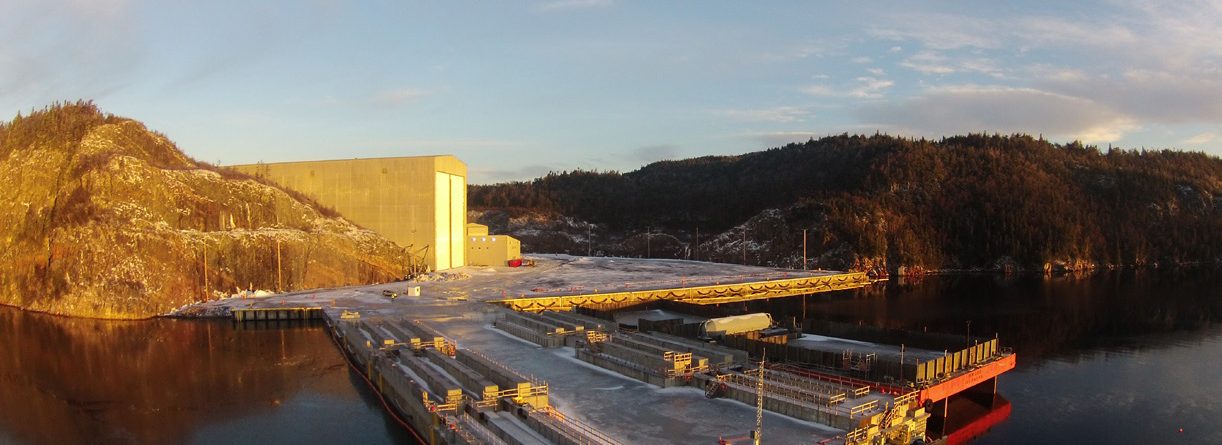 Aerial view of an industrial dock facility on a calm body of water, surrounded by forested hills and a large building under a clear sky with soft sunlight. Snow patches are visible on the ground and dock.
