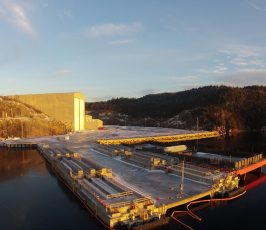 Aerial view of an industrial dock facility on a calm body of water, surrounded by forested hills and a large building under a clear sky with soft sunlight. Snow patches are visible on the ground and dock.