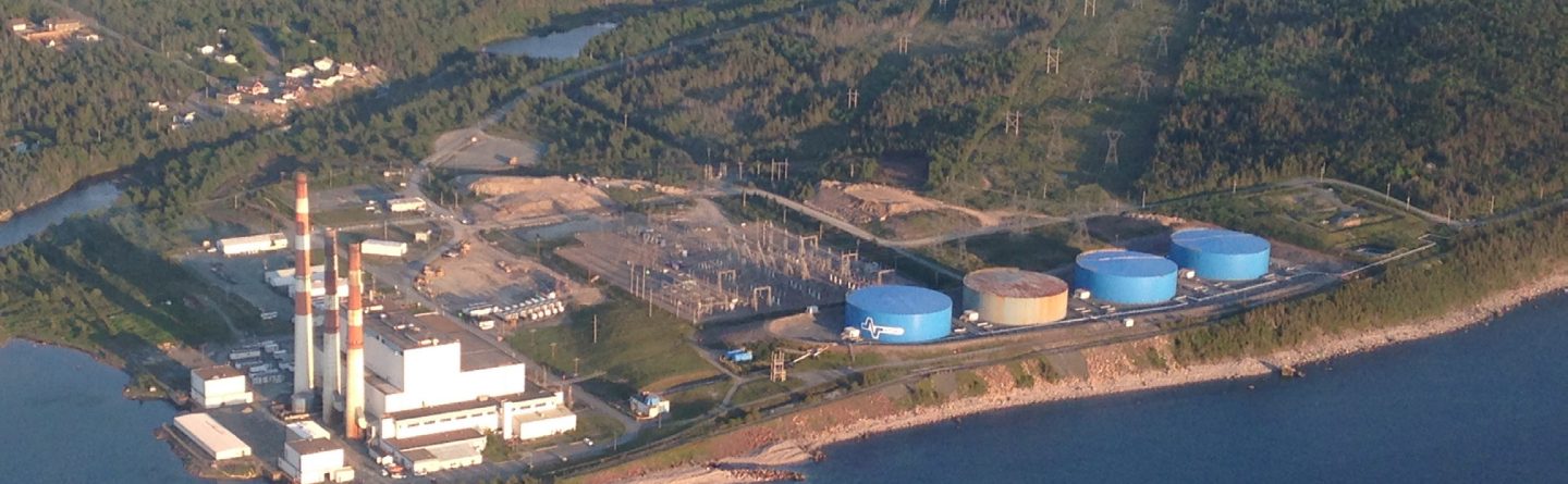 Aerial view of a coastal industrial facility with smoke stacks, white buildings, blue storage tanks, power lines, and surrounding forested area near water.