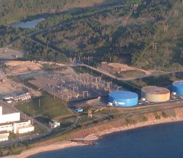 Aerial view of a coastal industrial facility with smoke stacks, white buildings, blue storage tanks, power lines, and surrounding forested area near water.