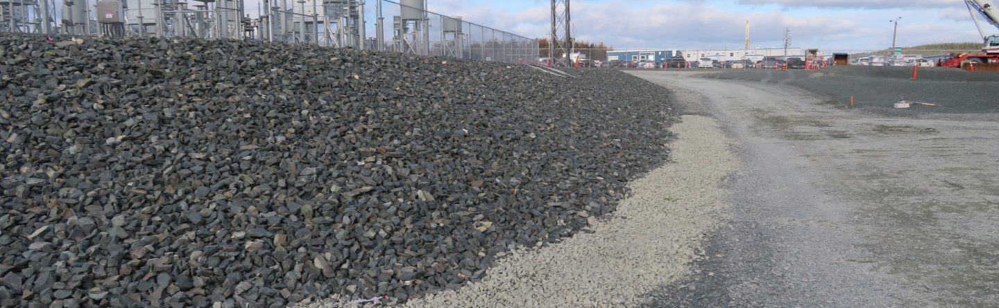 A gravel path runs beside a slope covered with larger rocks, next to an industrial facility with metal structures, fencing, and utility poles under a partly cloudy sky.