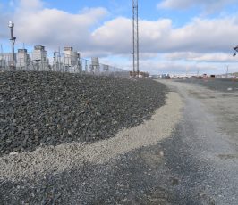 A gravel path runs beside a slope covered with larger rocks, next to an industrial facility with metal structures, fencing, and utility poles under a partly cloudy sky.