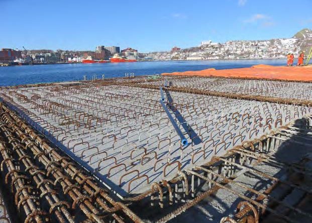 A large concrete slab with protruding steel reinforcement bars sits near a waterfront, with buildings and ships visible across the water under a clear blue sky. Two workers in orange clothing stand on the right.